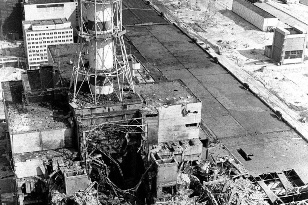 Foto aérea en blanco y negro de la central nuclear de Chernóbil en 1986: el techo y las paredes del reactor 4 están reventados, con hierros retorcidos y escombros bajo la alta chimenea a rayas.