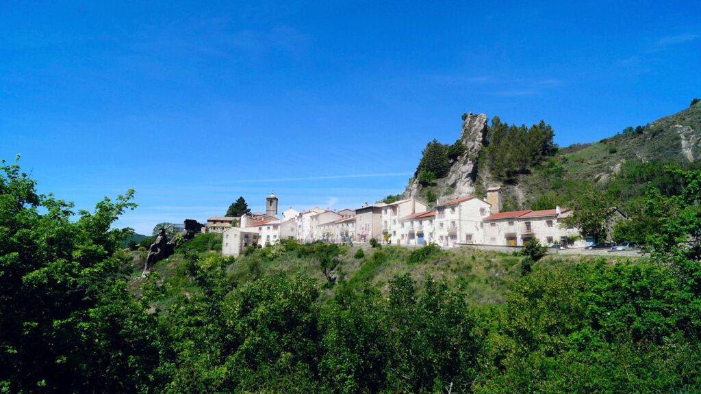 A small village of stone houses on a green hillside below a rocky crag, under a deep blue sky.