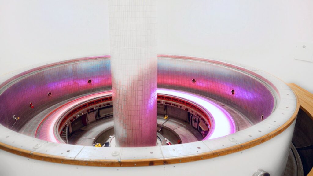 Vista del interior de una cámara de fusión tokamak en forma de rosquilla: un anillo de plasma rosa y violeta brilla alrededor de la columna central cubierta de placas y se refleja en las paredes metálicas.