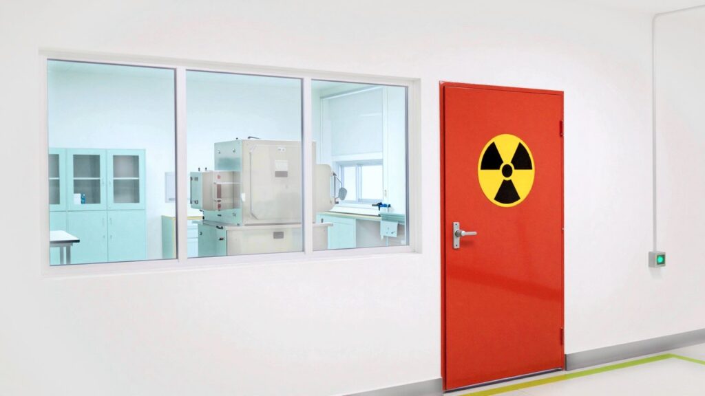 A sealed laboratory seen from a white corridor: large windows show steel glove boxes and turquoise cabinets inside, next to a closed coral-red door with a yellow and black radiation warning symbol.