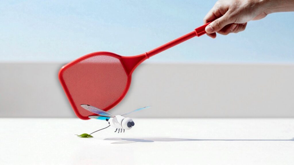 A hand swings a red flyswatter down at a tiny white robot with blue film wings hovering just above a white table, a split second before it hits.