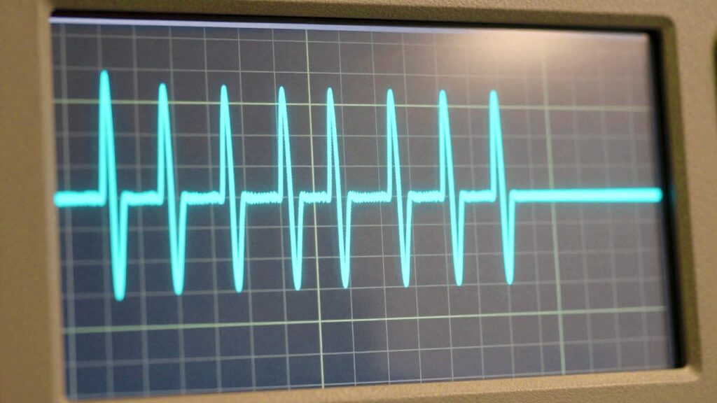 La pantalla de un osciloscopio con una onda sinusoidal brillante sobre una cuadrícula, en una mesa de laboratorio blanca.