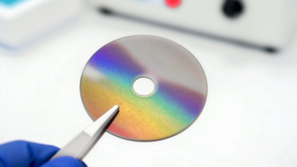 Un disco redondo sujeto con pinzas brilla con colores de arcoíris por una capa fina, sobre una mesa blanca.