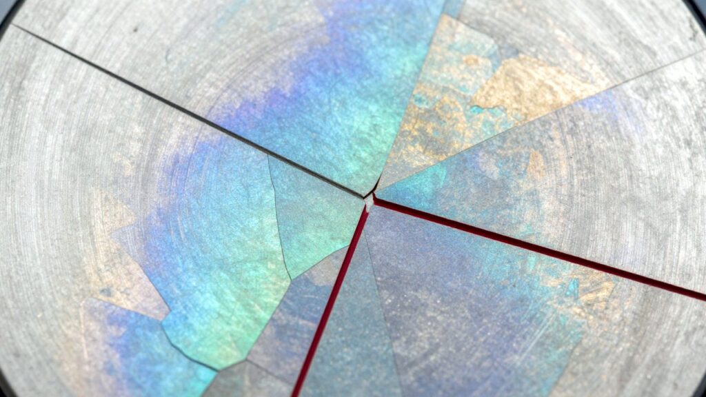 Primer plano de una muestra de metal pulida y atacada: granos cristalinos angulosos con brillos azules, turquesa, lilas y dorados, separados por finas líneas oscuras y rojas.