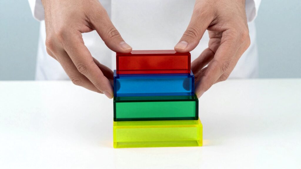 Two hands placing the top block on a staircase of glowing red, blue, green and yellow glass blocks on a white table.