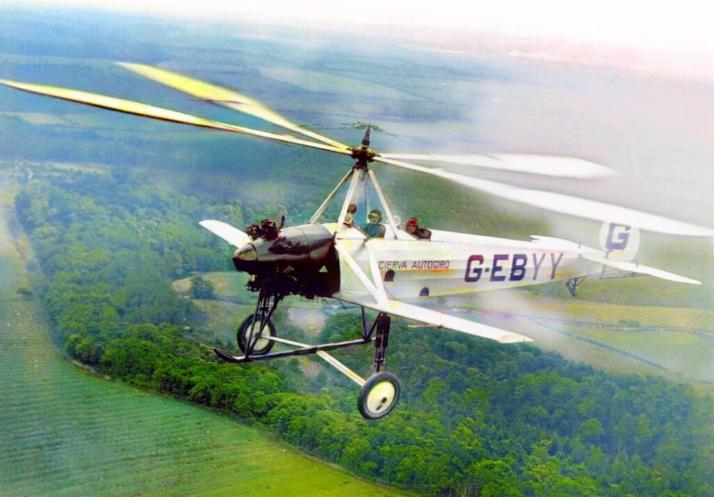Foto coloreada: un autogiro Cierva C.8L con matrícula G-EBYY vuela bajo sobre campos verdes y bosques, con las largas palas del rotor girando sobre el fuselaje.