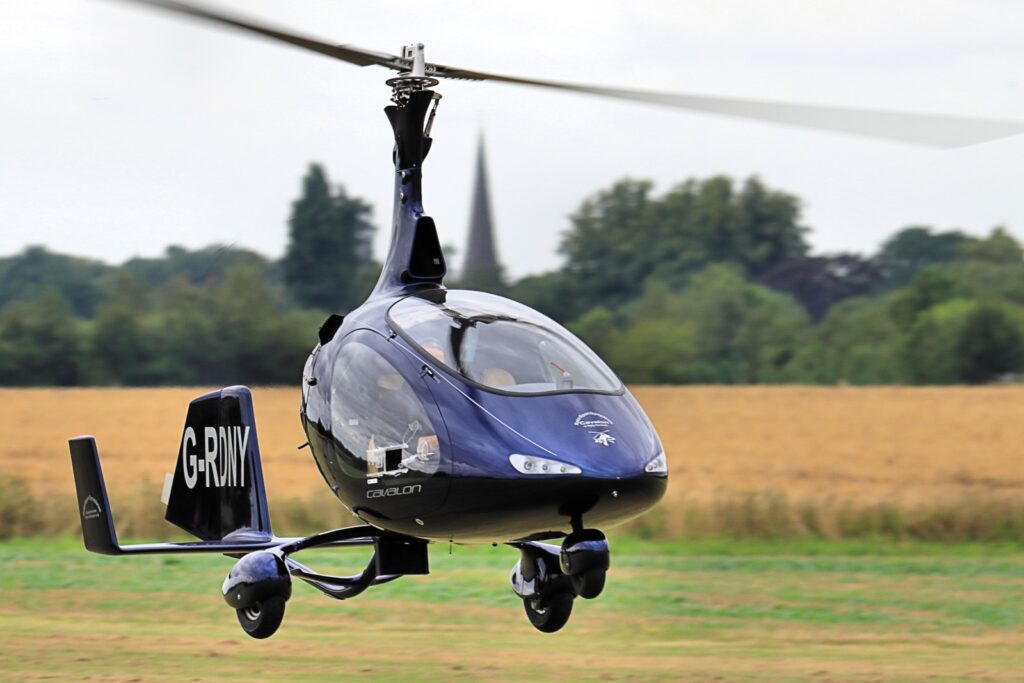 A modern blue AutoGyro Cavalon autogyro with an enclosed cockpit flies low over a golden field in front of green trees.